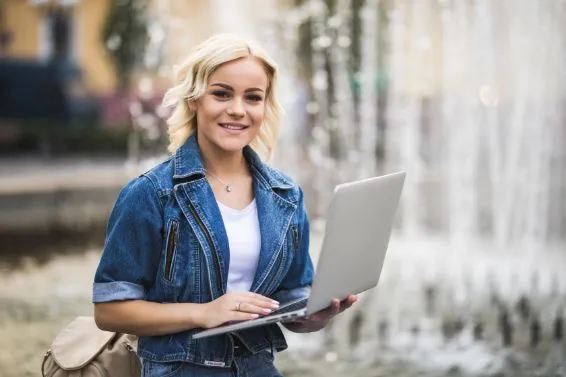 Eine Frau mit blonden, schulterlangen Haaren trägt eine Jeansjacke und hält einen Laptop in den Händen. Im Hintergrund sieht man einen Brunnen mit Wasserfontänen.